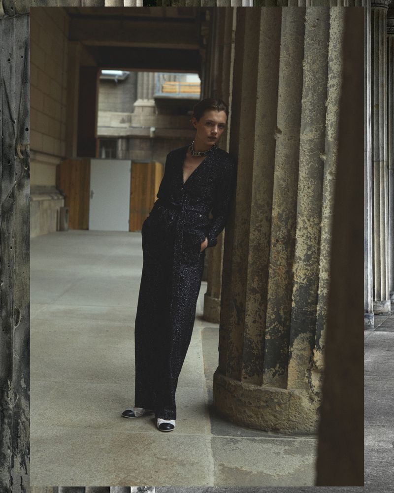 Fashion shot of a woman in a black jumpsuit leaning against a stone column outdoors in a courtyard of a building with stone archways.