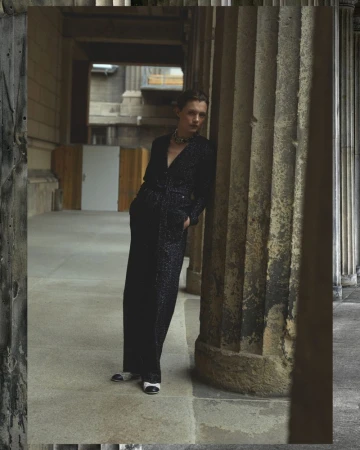 Fashion shot of a woman in a black jumpsuit leaning against a stone column outdoors in a courtyard of a building with stone archways.
