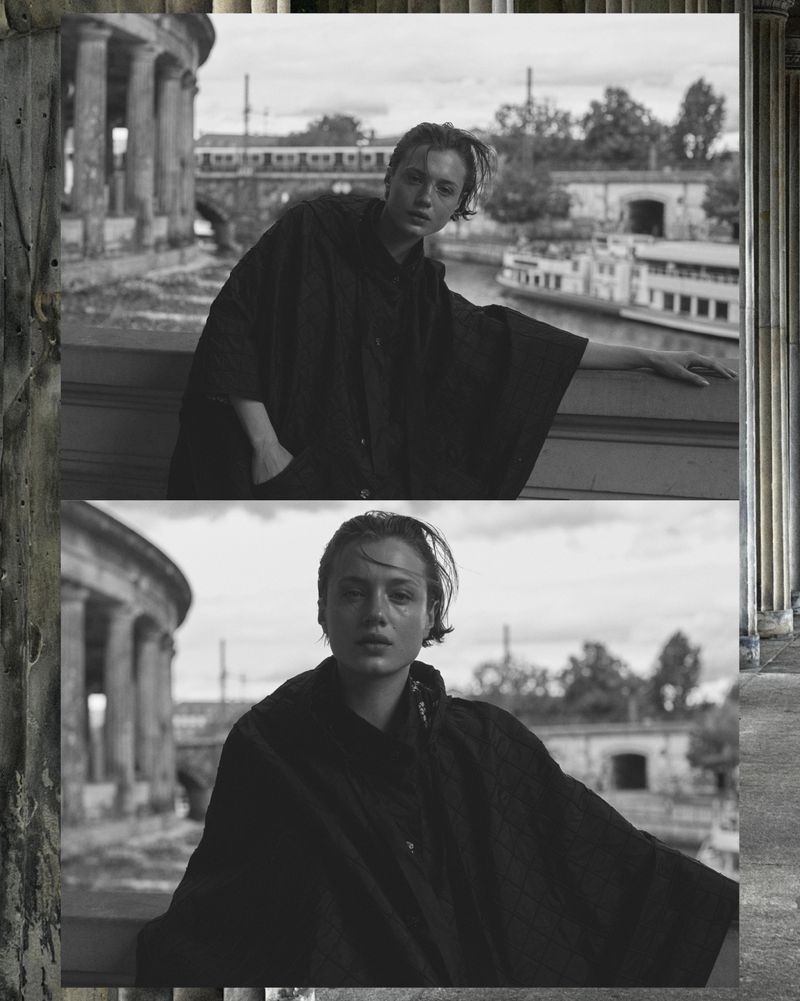 Monochrome portrait of a young adult woman in a black garment posing against a bridge, river, and colonnade backdrop in daylight.
