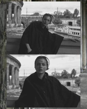 Monochrome portrait of a young adult woman in a black garment posing against a bridge, river, and colonnade backdrop in daylight.