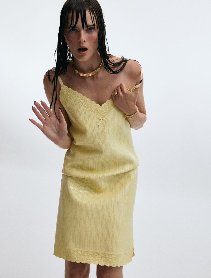 Model with wet-look bangs wearing a pale yellow lace-trimmed slip dress and gold choker necklace against a white background.