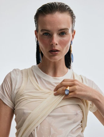 Model with slicked-back wet hair wearing a wet white t-shirt, blue stone drop earrings, and a large ring against a pale backdrop.