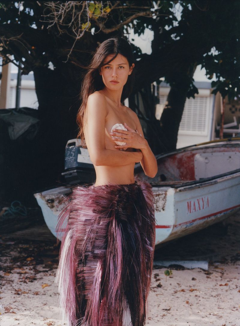 Mahalia Celeste Henderson wearing a fringed purple skirt and holding a shell, posing next to a boat on a sandy beach.