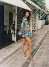 Model Mahalia Celeste Henderson standing barefoot outdoors in a floral bell-sleeve mini dress under a building awning.