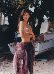 Mahalia Celeste Henderson wearing a fringed purple skirt and holding a shell, posing next to a boat on a sandy beach.