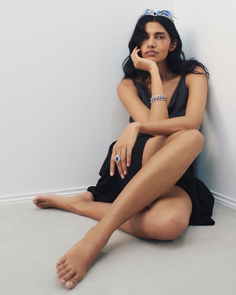 Model Pooja Mor seated on a white floor against a plain white corner wall, wearing a dark dress and statement jewel tiara.