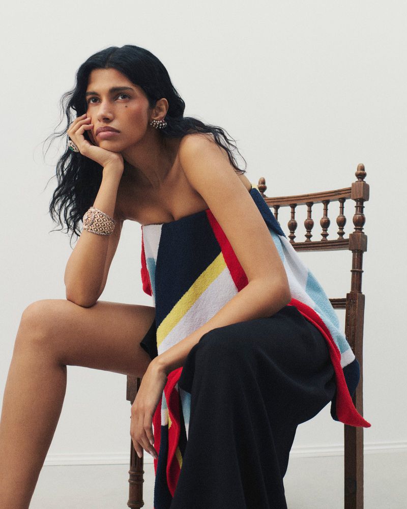 Model Pooja Mor seated on a wooden spindle chair with her chin in hand, wearing a geometric wrap, black trousers, and statement jewelry.