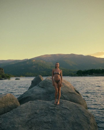 Woman wearing a two-piece bikini standing atop a large boulder in a lake with mountains in the background.