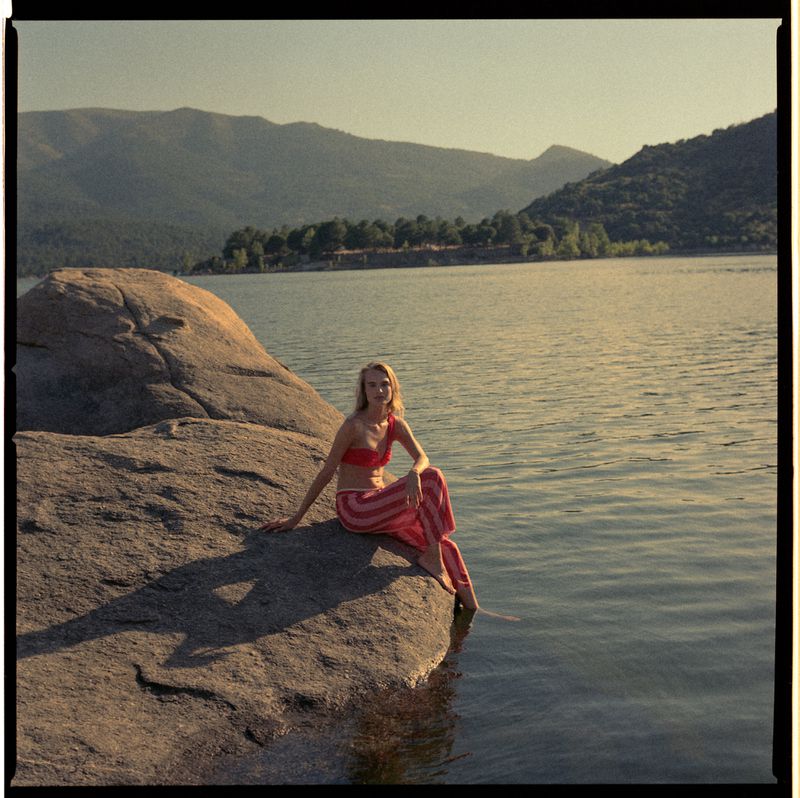 Woman in a red bikini top and striped pink pants sitting on a large boulder by a calm lake with mountains in the background.