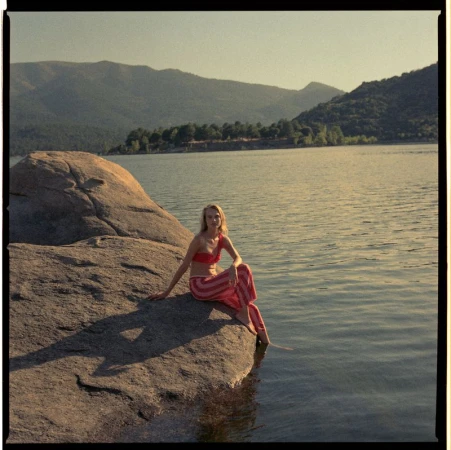 Woman in a red bikini top and striped pink pants sitting on a large boulder by a calm lake with mountains in the background.