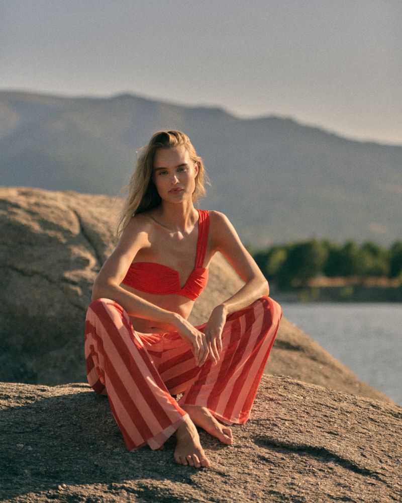 A woman in a red one-shoulder top and red and pink striped trousers sits barefoot on a large boulder with hills in the background.