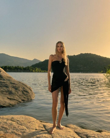 Blonde woman standing barefoot on rocky lakeshore wearing a black one-shoulder swimsuit and sheer black wrap.