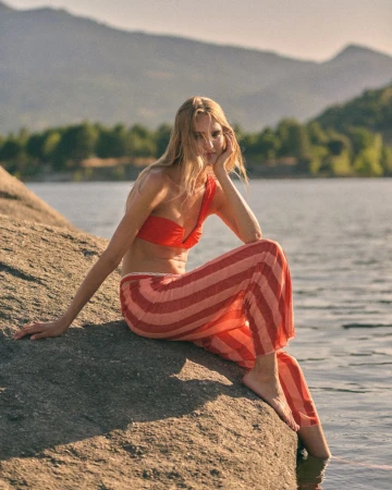 Woman in a red one-shoulder top and striped pink trousers sitting on a sunlit rock beside a calm lake with mountains.