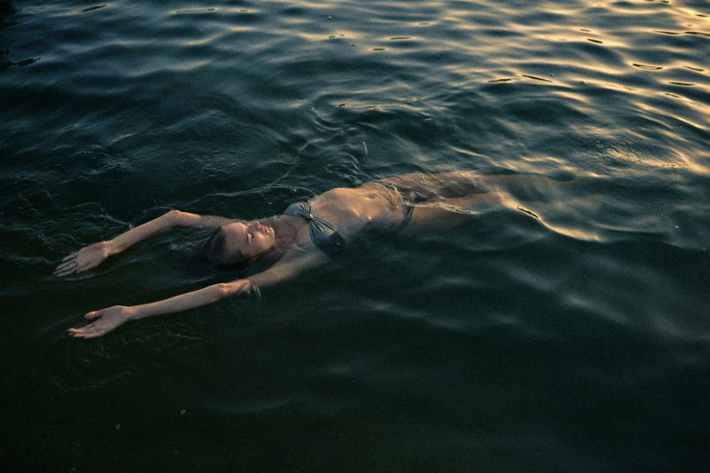 Woman floating on her back in dark rippling water wearing a two-piece swimsuit with golden light reflecting across the surface.