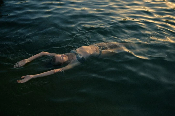 Woman floating on her back in dark rippling water wearing a two-piece swimsuit with golden light reflecting across the surface.