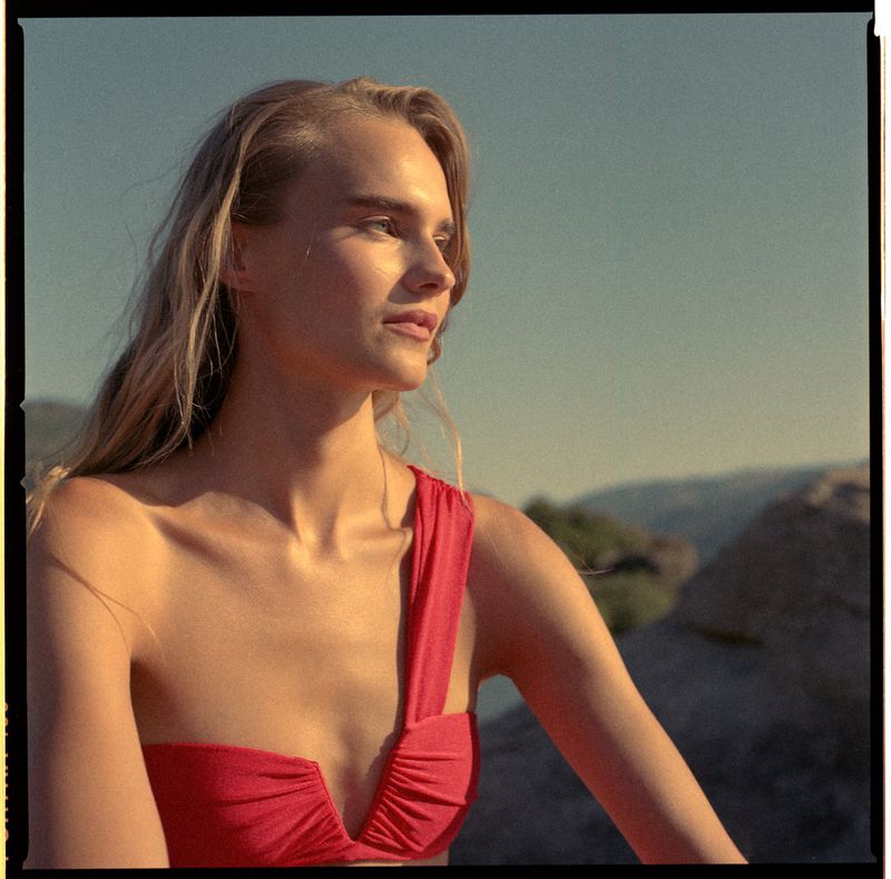 Woman in a red one-shoulder swimwear top outdoors in warm sunlight against a rocky coastal landscape background.
