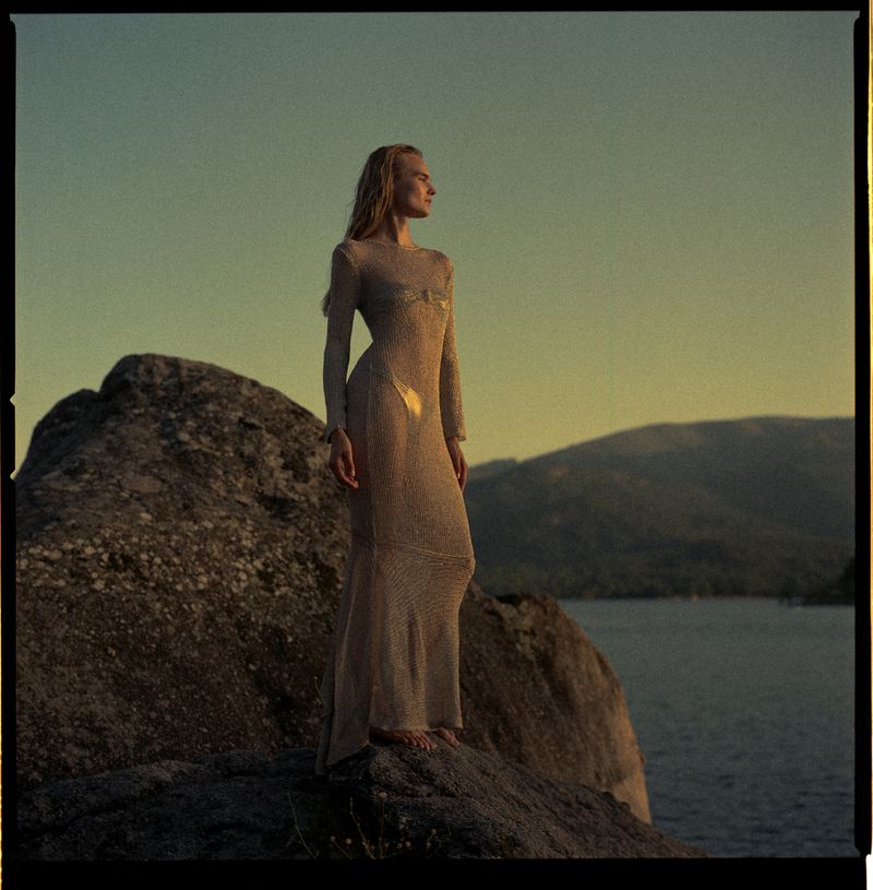 Woman in a long sheer dress standing on a rocky outcrop overlooking a lake at sunset
