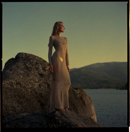 Woman in a long sheer dress standing on a rocky outcrop overlooking a lake at sunset