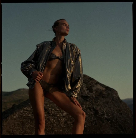 Woman posing on a rock in a dark metallic bikini and oversized iridescent bomber jacket under evening sky.