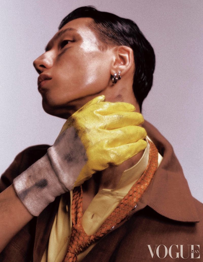 Profile of an East Asian man wearing a brown jacket while a hand in a distressed yellow work glove touches his neck.