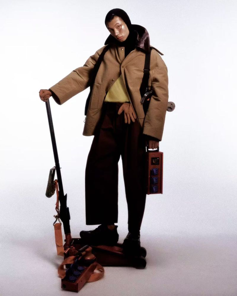 An Asian male model standing on studio equipment wearing a tan jacket, dark trousers, and a black balaclava against a white background.