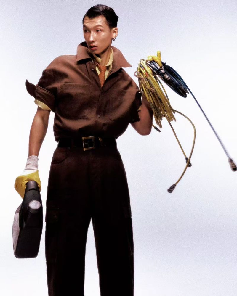 Asian male model in a brown workwear uniform holding an oil jug and tangled cables against a white background.