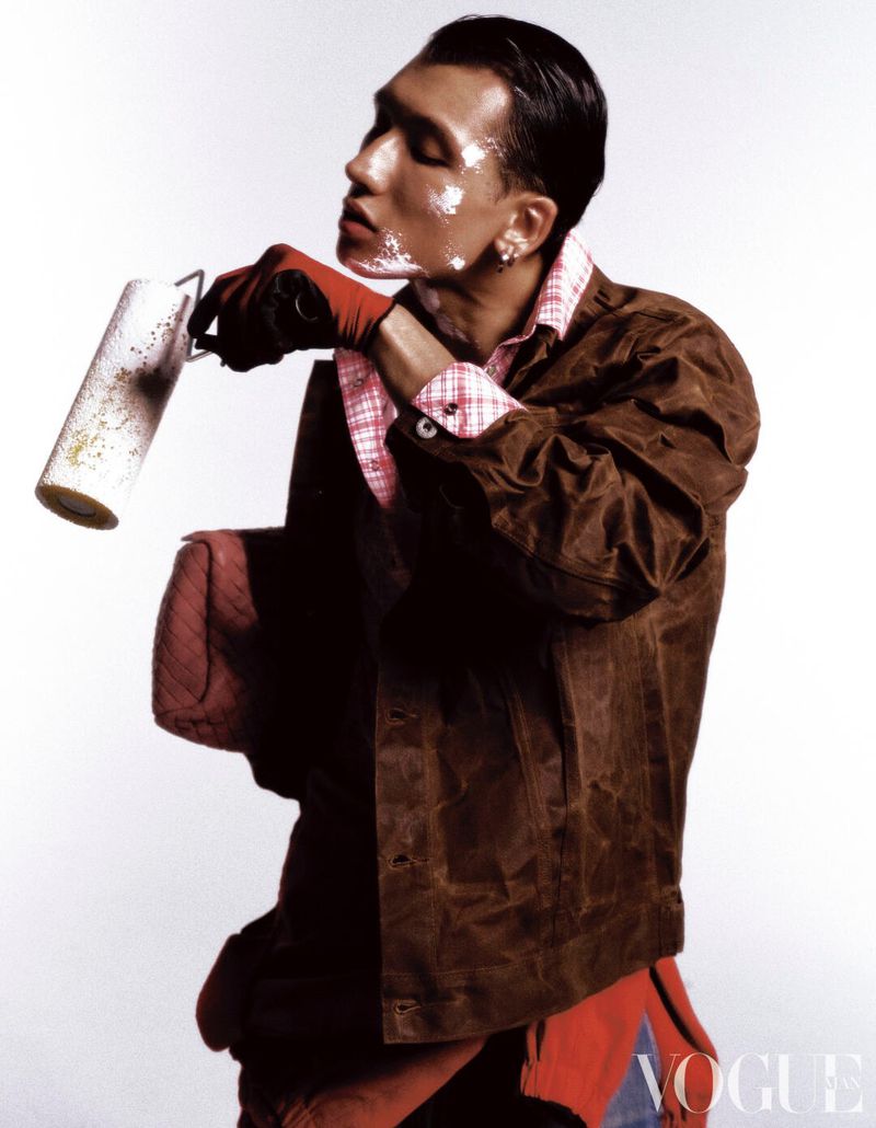 Model with white paint markings on his face holding a paint roller while wearing a brown workwear jacket on a white backdrop.