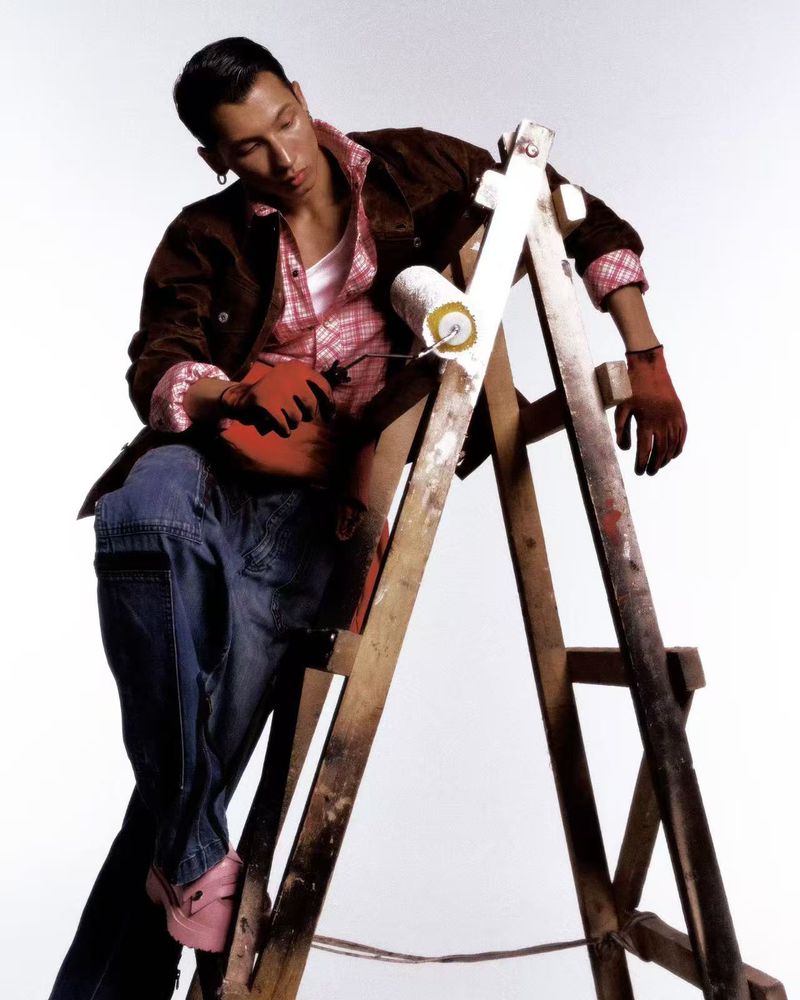 East Asian male model in a brown jacket and denim jeans seated on a paint-splattered wooden ladder holding a paint roller.
