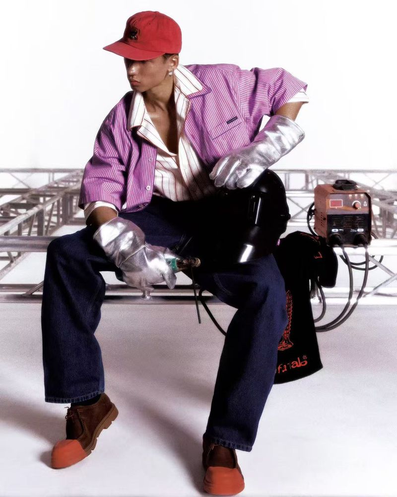 Model wearing a pink striped shirt, red baseball cap, and silver protective gloves sitting against a white seamless backdrop.