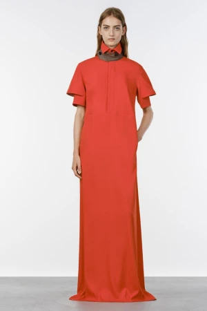 Model wearing a bright coral red short-sleeved floor-length dress with a high collar against a plain studio background.