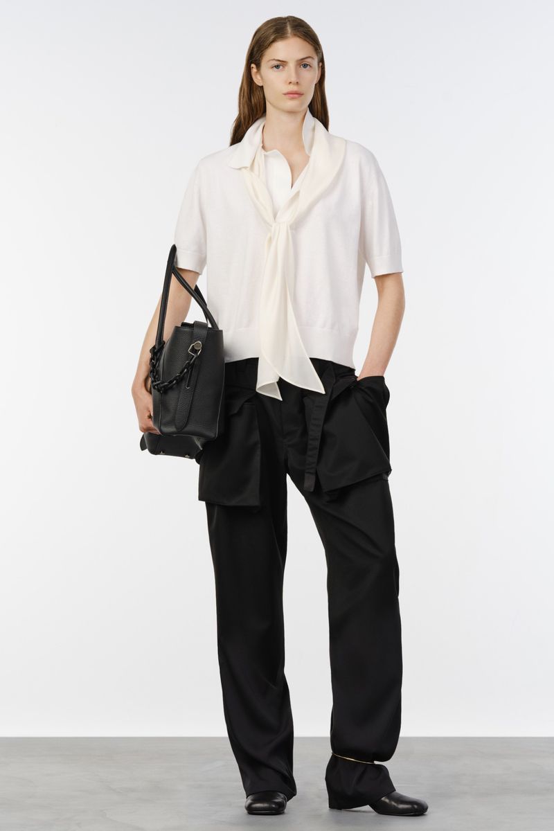 Model posing in a cream short-sleeve draped top, relaxed black utilitarian trousers, and black leather bag for Sacai.