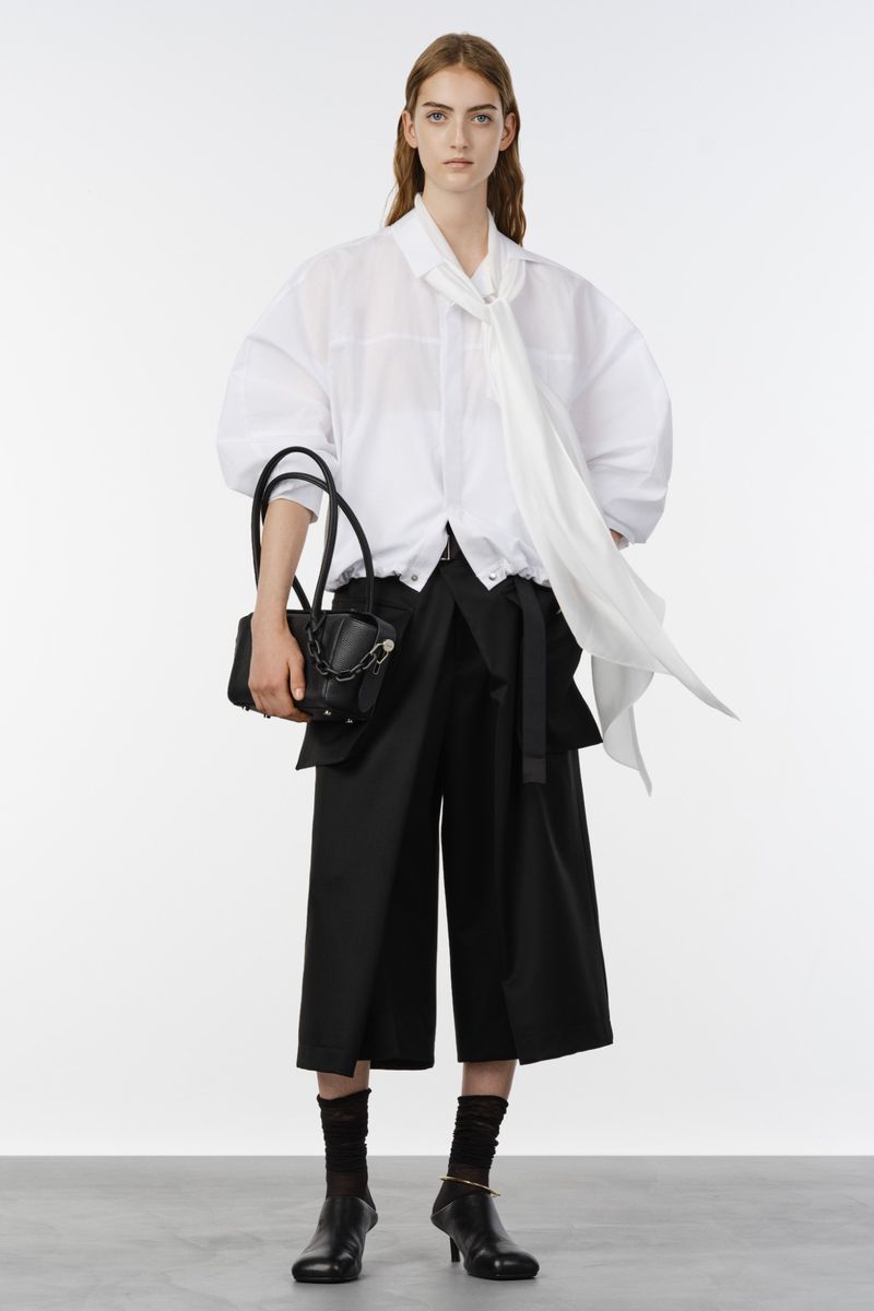 Model posing in an oversized white tie-neck blouse, black culottes, and low heels with a black handbag against a studio backdrop.