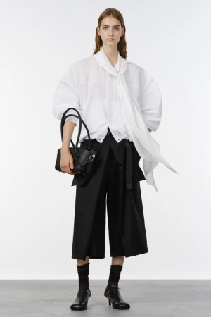 Model posing in an oversized white tie-neck blouse, black culottes, and low heels with a black handbag against a studio backdrop.