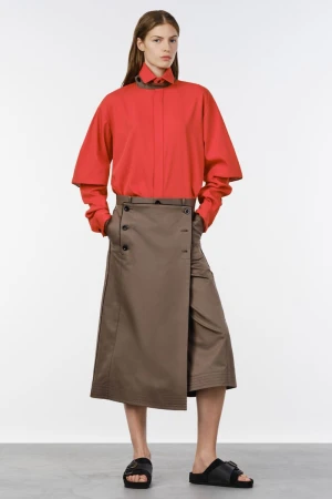 Model wearing a bright red collared shirt and an asymmetric taupe wrap skirt with black slide sandals in a studio setting.