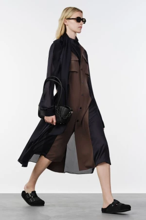 Model walking wearing sunglasses, a layered brown and black coat dress, black mule shoes, and holding a black leather handbag.