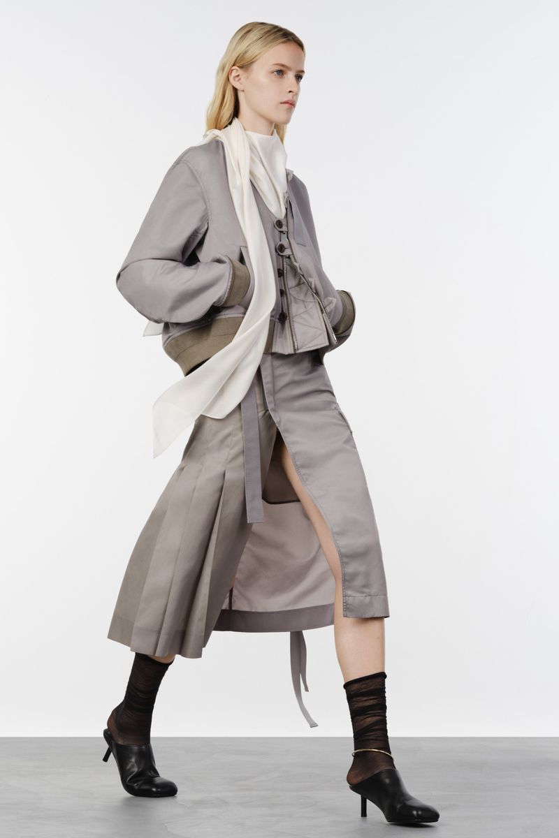 Model walking in a grey layered jacket, slit pleated skirt, flowing white scarf, and black heels on a white studio background.