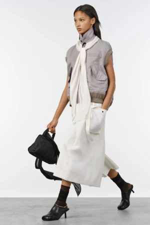 Model in motion wearing a grey utility vest, white draped scarf, white skirt, and black mules against a white background.