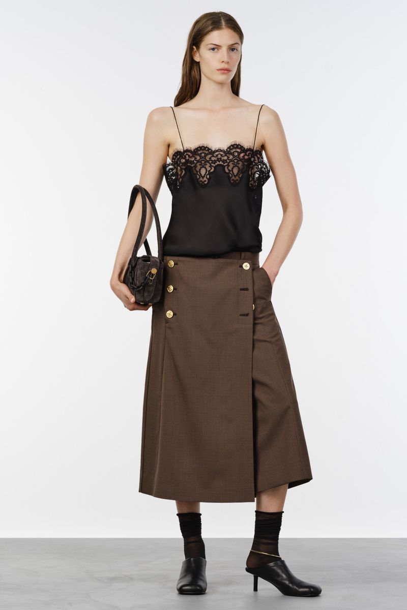 Model wearing a black lace-trim camisole and brown button-front midi skirt holding a dark handbag against a white studio background.