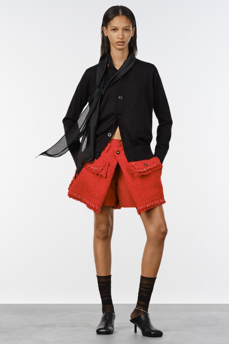 Model Elodie Guipaud wearing a black buttoned cardigan with scarf and a textured red mini skirt on a white studio background.