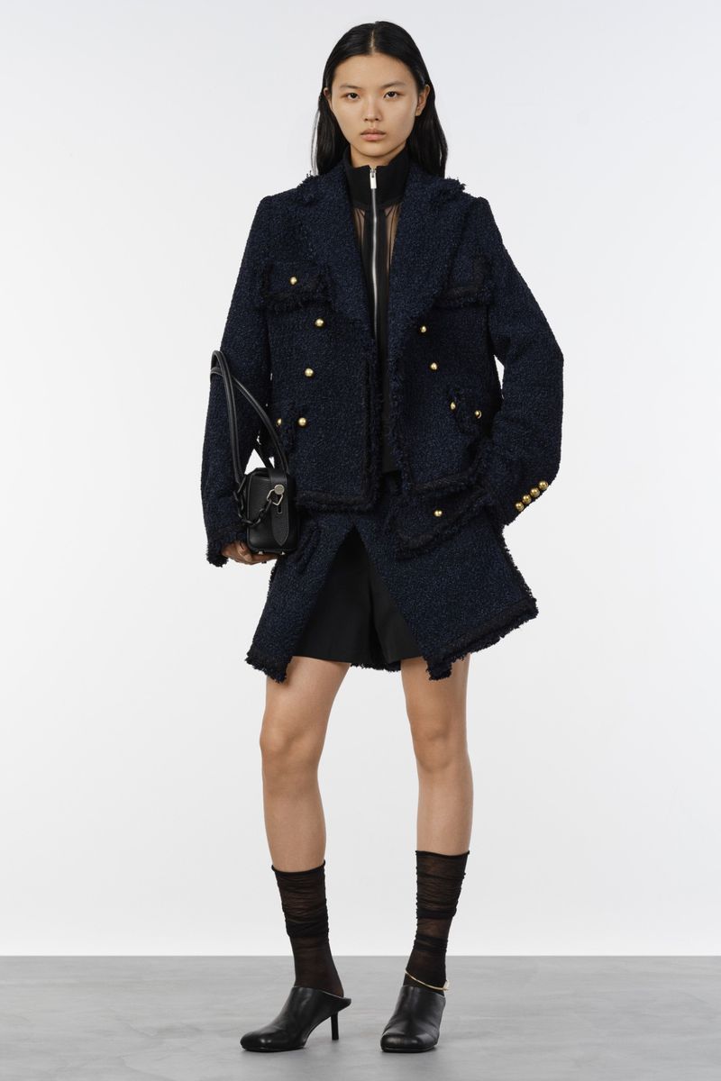 Model standing against a white backdrop wearing a dark blue tweed jacket, black shorts, sheer socks, and black heels while holding a handbag
