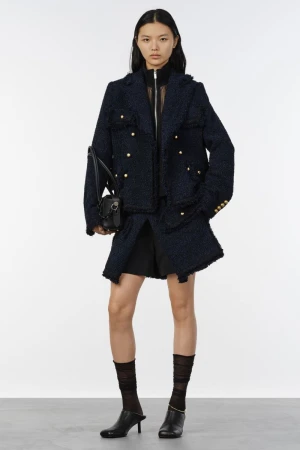 Model standing against a white backdrop wearing a dark blue tweed jacket, black shorts, sheer socks, and black heels while holding a handbag