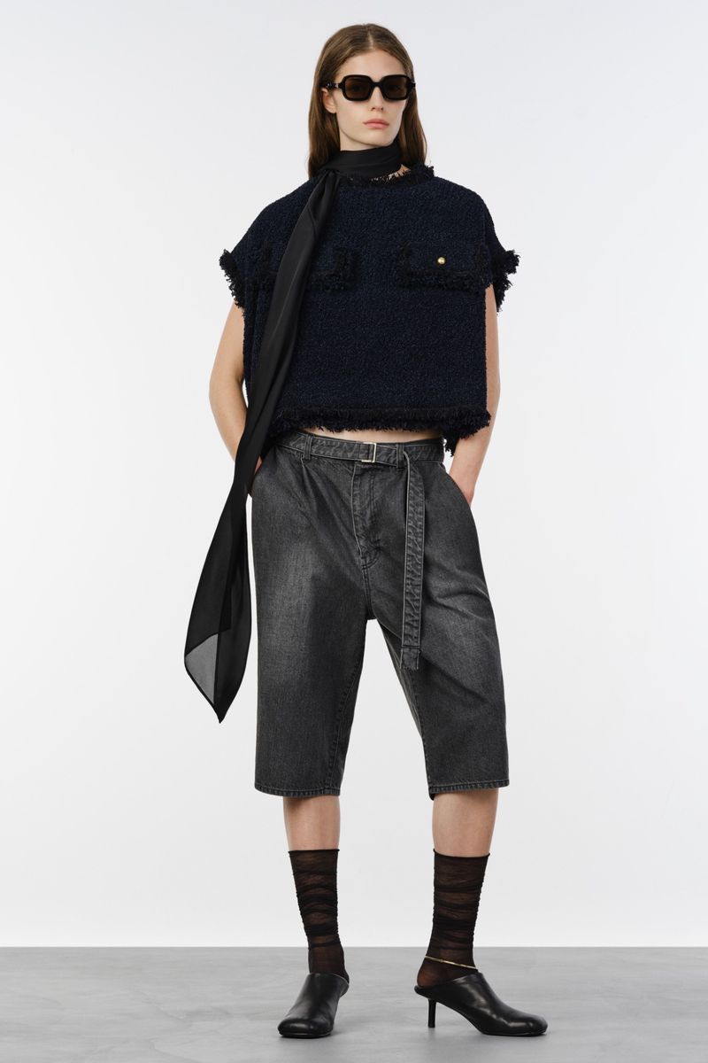 Model wearing dark sunglasses, a cropped dark tweed top, belted grey denim shorts, and black kitten heels against a white background.