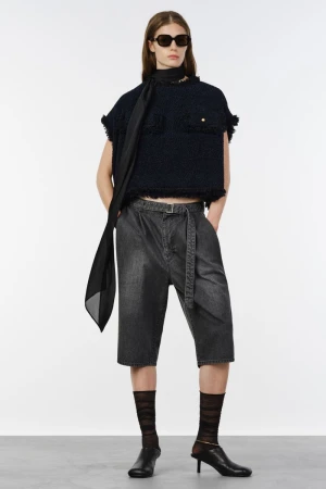 Model wearing dark sunglasses, a cropped dark tweed top, belted grey denim shorts, and black kitten heels against a white background.