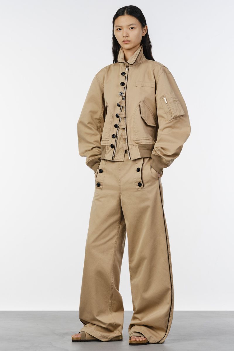 Model wearing a tan bomber jacket and wide-leg trousers against a white seamless background for Sacai