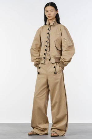 Model wearing a tan bomber jacket and wide-leg trousers against a white seamless background for Sacai