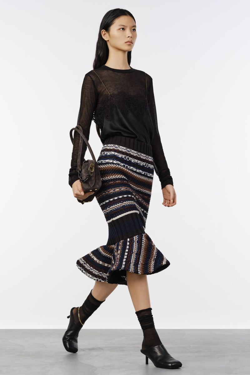 Model walking in a black sheer top and patterned knit skirt carrying a brown handbag against a grey backdrop