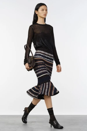 Model walking in a black sheer top and patterned knit skirt carrying a brown handbag against a grey backdrop
