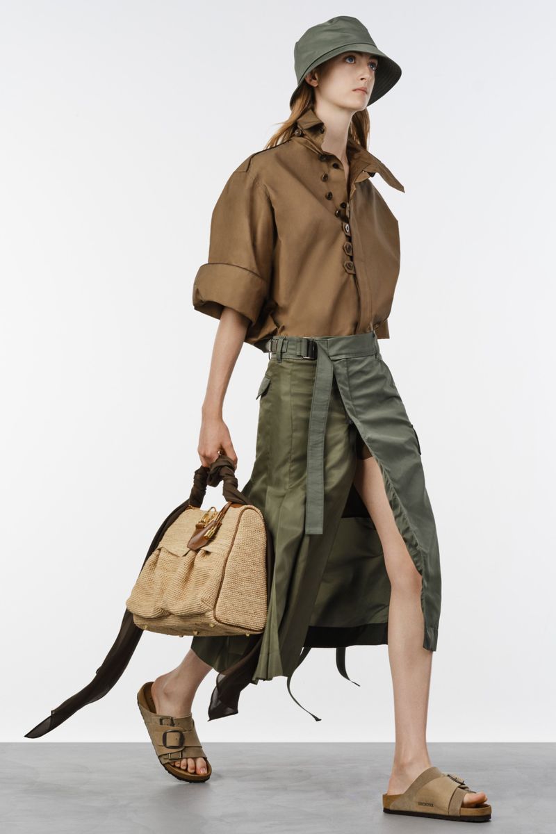 Model walking in a brown short-sleeve shirt, green wrap skirt, matching bucket hat, and sandals holding a beige bag against a white backdrop.