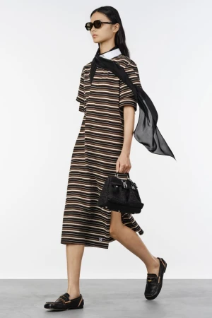 Model in a striped midi polo dress with sunglasses, a flowing black scarf, and loafers against a white studio background