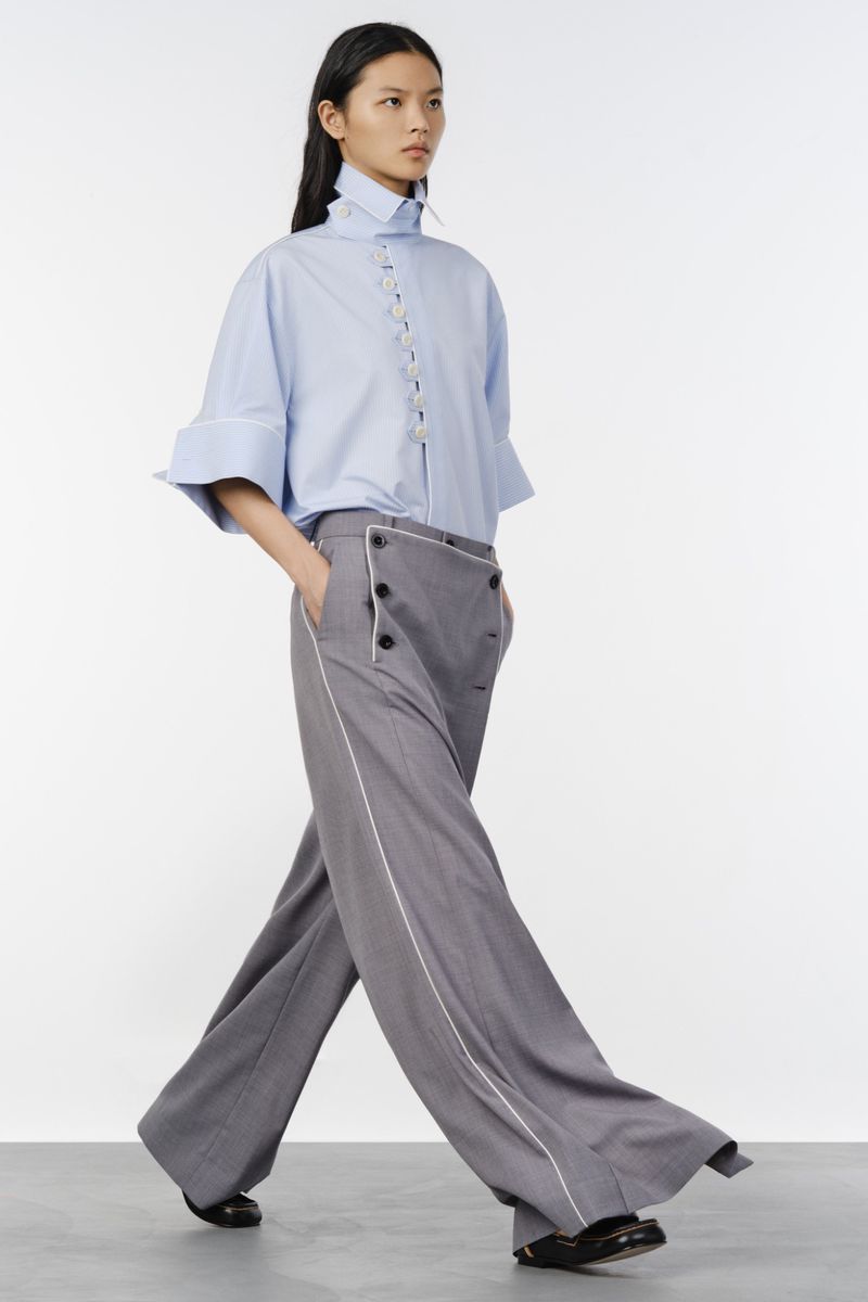 Fashion model walking in a light blue short-sleeve shirt and high-waisted wide-leg grey trousers against a white backdrop.